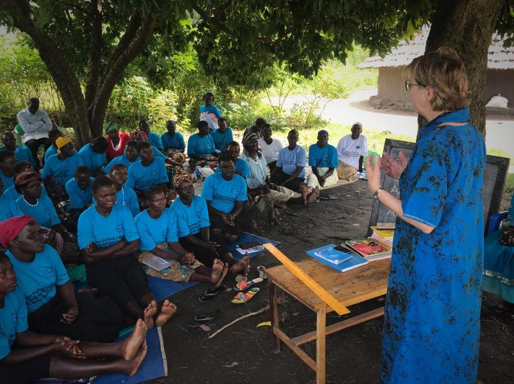 Karon standing, speaking to group of seated Ugandan women outdoors.